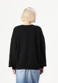 Fekete, oversized kapucnis pulóver, ejtett vállakkal, bordázott mandzsettákkal és texturált felülettel. Világoskék farmernadrág felett viselve.