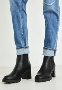 Botas de cuero negras hasta el tobillo con un tacón grueso y paneles laterales elásticos. Llevadas con jeans de mezclilla azul claro, doblados. Textura de superficie suave.