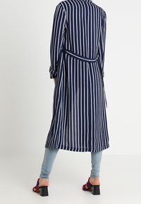 Abito lungo a righe navy e bianche con cintura. Presenta maniche con bottoni e un giromanica sul retro. Indossato sopra jeans azzurri e sandali con tacco.