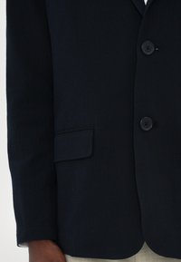 Navy blazer with a textured finish, two buttons, and a single chest pocket. Sleeves are long with a straight cut, complementing formal attire.