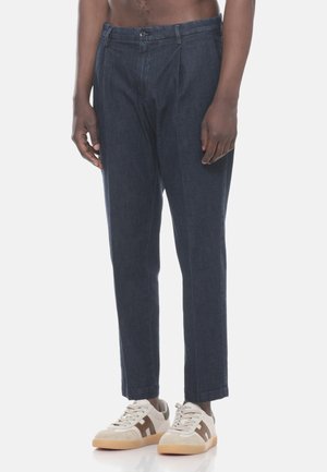 Uomo che indossa pantaloni dritti blu scuro e sneakers beige con dettagli marroni, in piedi davanti a uno sfondo bianco semplice.