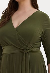 Ελαιώδη πράσινο περιτύλιγμα φόρεμα από μαλακό ύφασμα, με βαθύ V λαιμόκοψη και κόμπο στη ζώνη, με τρία τέταρτα μανίκια.