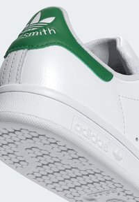 Hvit skinnsneaker med grønn hælflik, teksturert såle og "adidas"-merke. Lave sko, glatt overflate og syede detaljer.