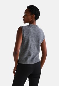 Gilet sans manches en tricot gris avec un col et un ourlet côtelés, présentant une texture subtile. Porté sur un pantalon noir, vu de dos.