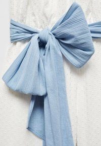 Ljusblått tygbälte knutet i en stor rosett, tillverkat av mjukt, strukturerat material, bärs över ett vitt plagg med prickigt mönster.