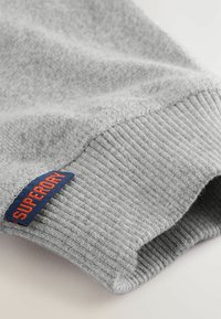 Grijze sweatstoffabric met een gestructureerd oppervlak, voorzien van een geribde manchetkant en een blauw label met de tekst "SUPERDRY" in oranje letters.