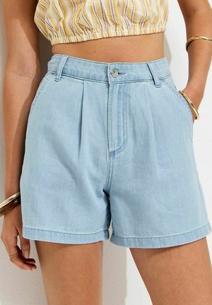 Shorts di jeans azzurri chiaro a vita alta con pieghe, indossati con un crop top giallo a righe, accessoriati con bracciali e anelli dorati.