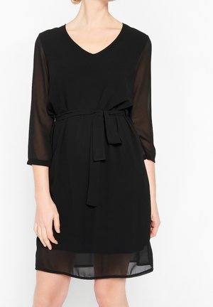 Woman wearing a knee-length black dress with a V-neck, sheer three-quarter sleeves, and a tied fabric belt at the waist.