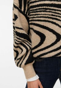 Béžový pletený svetr s černými vlnkovými vzory, mírně oversized střih a žebrované manžety. Nošený s tmavými džínami a bílou košilí pod ním.