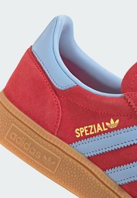 Červené semišové tenisky Adidas so svetlomodrými pruhmi a golierom, žltý text "SPEZIAL" a gumená podrážka s logom Adidas na strane.