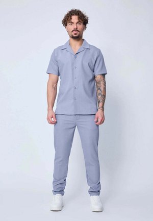 Homme se tenant face à l'avant, portant une chemise à manches courtes bleu clair avec des boutons, un pantalon assorti, des baskets blanches et un tatouage détaillé sur le bras.