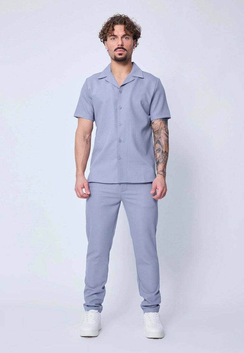 Homme se tenant face à l'avant, portant une chemise à manches courtes bleu clair avec des boutons, un pantalon assorti, des baskets blanches et un tatouage détaillé sur le bras.