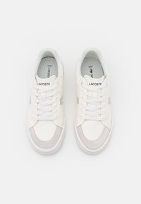 Chaussures de tennis blanches avec une tige texturée et des accents en daim gris. Présentent des lacets plats, un embout arrondi et un logo sur la languette.