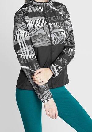 Kvinde iført sort og hvid mønstret jakke med lynlås og abstrakte designs samt turkise leggings, hænder krydset ved livet.