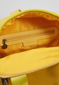 Interior de bolsa de tela amarilla con un revestimiento suave. Presenta un bolsillo con cremallera y una etiqueta de marca. Diseño simple, que enfatiza la practicidad y la organización.