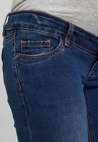 Primer plano de una persona que lleva puesto un pantalón de mezclilla azul con costuras naranjas y una camisa gris metida en la cintura.