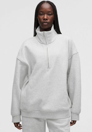 Donna con i capelli intrecciati che indossa una felpa grigio chiaro con mezza zip e pantaloni abbinati, in piedi davanti a uno sfondo neutro semplice.