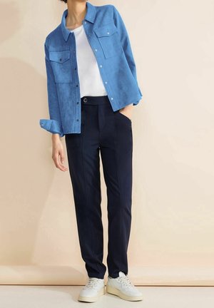 Person iført en blå skjorte med knapper over en hvid top, sorte bukser og hvide sneakers, der står mod en beige baggrund.