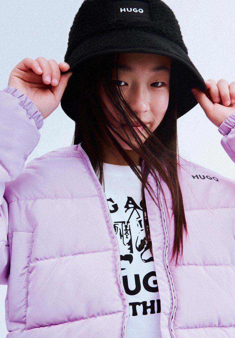 Lila pufferjack met een quilted ontwerp en geborduurd "HUGO" logo. Zwarte fluffy bucket hat met logo aan de voorkant. Witte graphic t-shirt eronder.