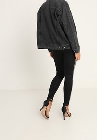 Zwarte denimjack met een relaxed fit, gecombineerd met zwarte leggings met zijstrepen en zwarte hoge hakken. Zachte texturen zichtbaar.