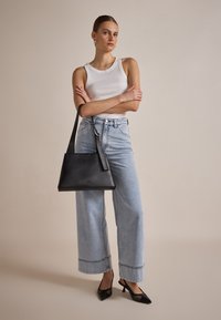 Schwarze Lederhandtasche mit strukturierter Form, flachem Boden und Schultergurt. Das Model trägt ein weißes Tanktop und hellblaue weit geschnittene Jeans.