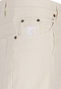 Beige bukser i et robust stof har en baglomme med en hvid broderet logo og messing hardware detaljer. Synlige sømme langs siderne.