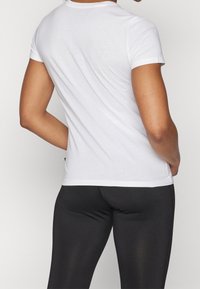 Camiseta blanca de algodón con mangas cortas, cuello redondo y un diseño ajustado, llevada con leggings negros, con costuras visibles en el dobladillo.