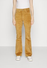 Corduroy weit geschnittene Hose in Senfgelb, mit hoher Taille, frontalen Taschen und vertikaler Ripptextur. Kombiniert mit weißen Sneakers.