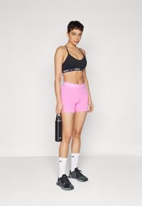 Černá sportovní podprsenka s logem Nike, růžové vysoké šortky, černé tenisky a bílé ponožky s malými logy. Drží černou láhev na vodu.