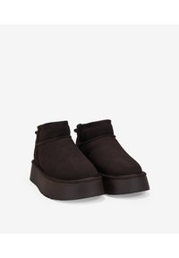 Botas de tobillo de ante marrón oscuro con suela de plataforma gruesa, puntera redondeada y costuras mínimas a lo largo de las uniones. Sin hardware ni patrones visibles.