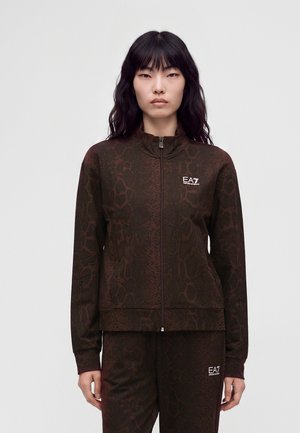 Femme aux longs cheveux foncés porte une veste zippée marron foncé à motif et un pantalon assorti avec des logos blancs EA7.