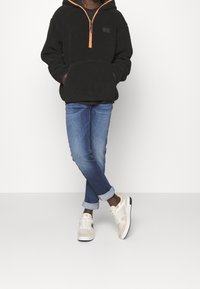 Felpa nera in felpa con cappuccio e zip arancione, tasca frontale e patch logo; abbinata a jeans in denim blu e sneakers beige con dettagli neri.