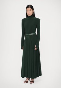 Tumši zaļa kleita ar garām piedurknēm, augstu apkakli, plisētu svārku un atbilstošu jostu viduklī. Gluda auduma ar plūstošu dizainu.