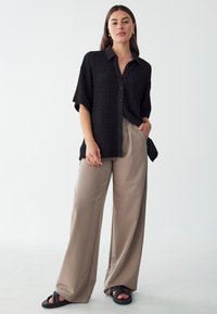 Camisa negra de textura con botones y mangas cortas, combinada con pantalones de pierna ancha en beige claro y sandalias negras de tiras.