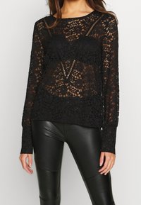 Haut à manches longues en dentelle noire avec un motif floral, une texture transparente et un décolleté en V. Assorti à des leggings ajustés en simili cuir noir.
