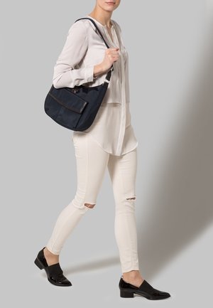 Laivastonsininen pehmeäntuntuinen käsilaukku, jossa on edessä tasku ja ruskeat yksityiskohdat. Yhdistetty kevyeen, vaaleaan paitapuseroon ja vaaleanbeigeihin revittyihin farkkuihin.