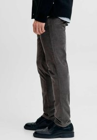 Pantalones de pana marrones con canalones verticales, diseño ajustado y sutiles detalles de costura roja, combinados con zapatos negros de cordones.