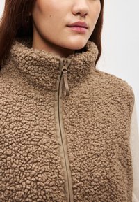 Καφέ γιλέκο από φλις με υψηλό γιακά, υφή επιφάνειας και κλείσιμο με φερμουάρ μπροστά, με αντίθετα λείες πάνελ στο φερμουάρ.