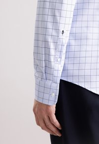 Camicia azzurra chiara con motivo a quadri navy, dotata di colletto standard, polsini abbottonati e un delicato ricamo sulla manica.