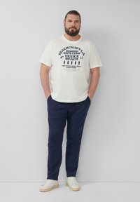 Witte katoenen T-shirt met marineblauwe grafische print; gecombineerd met marineblauwe broek en witte sneakers. Model staat tegen een effen achtergrond.