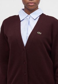 Personne portant un cardigan marron foncé boutonné par-dessus une chemise bleu clair à col, avec un petit logo crocodile vert sur le cardigan.