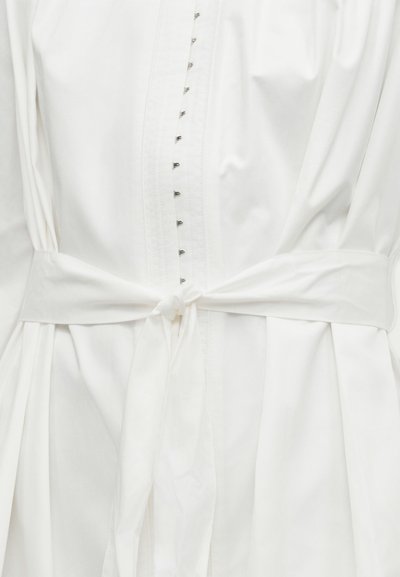 Λευκό ρούχο με μακριά μανίκια, κεντρική σειρά μικρών γαντζών και φαρδιά υφασμάτινη ζώνη δεμένη κόμπο στη μέση.