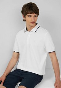 Weißes Poloshirt mit navyfarbenem Kragen und einer Drei-Knopf-Leiste. Leichte Textur, besticktes Logo auf der Brust, kombiniert mit navyfarbenen Shorts.