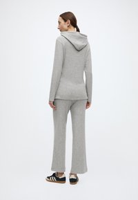 Pelēks džemperis ar kapuci un atbilstošas pelēkas bikses, abas izgatavotas no mīksta, teksturēta auduma. Apģērbu papildina melni baltas kedas.