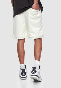 Shorts de sport en mesh blanc avec poches latérales, dotés d'une coupe ample et d'un ourlet roulé. Portés avec des baskets noir et blanc et des chaussettes blanches.