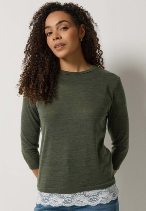 Vrouw met krullend haar, gekleed in een olijfgroene top met lange mouwen en een witte kanten zoom, en een blauwe spijkerbroek, staand tegen een lichtgrijze achtergrond.