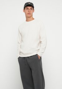 Sudadera texturizada color beige claro con puños acanalados, combinada con pantalones de chándal de corte relajado color gris oscuro. El modelo lleva una gorra oscura.