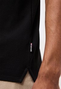 Gros plan sur l'ourlet texturé d'une chemise noire avec une petite étiquette de marque Schott et une partie d'un bras tatoué visible.