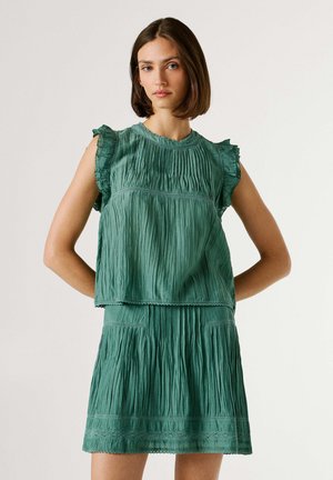 Jonge vrouw met bruin haar op schouderlengte, gekleed in een mouwloze groene top met structuur en een bijpassende rok, tegen een effen achtergrond.