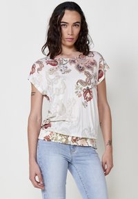 Haut à manches courtes de couleur crème avec des motifs paisley floraux rouges et verts intricats, présentant un ourlet inférieur froncé et une coupe décontractée.